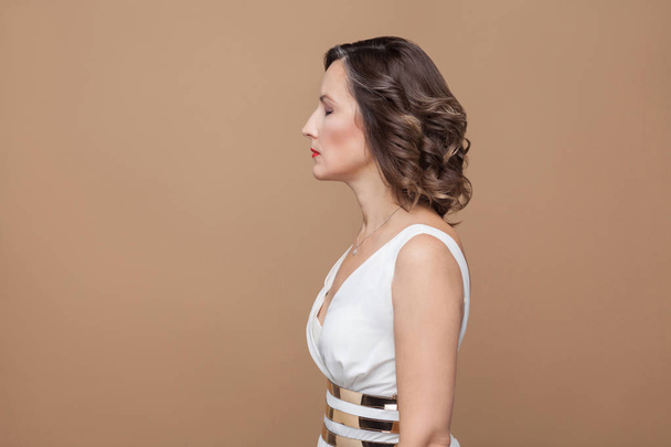  side view of sad woman with closed eyes in white dress with red lips and dark curly hairstyle posing on brown background - Photo, image