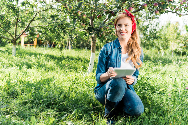 привлекательный фермер присел и держит планшет в яблоневом саду на ферме
 - Фото, изображение