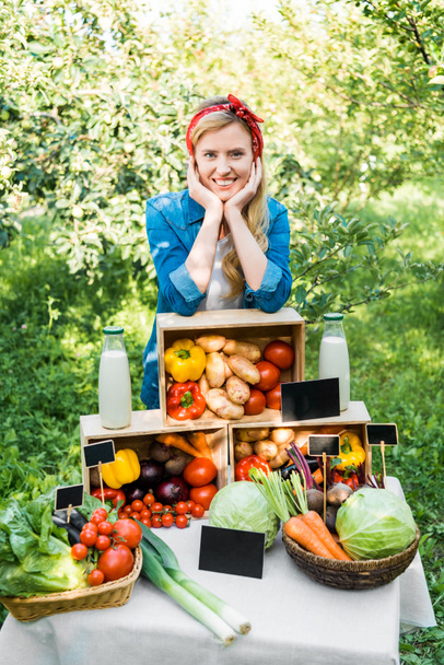 smiling farmer leaning on boxes with vegetables at farmer market - Фото, изображение