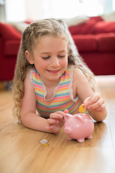 Young Girl Lying On Floor At Home Putting Coins Into Piggy Bank - Foto, Imagen