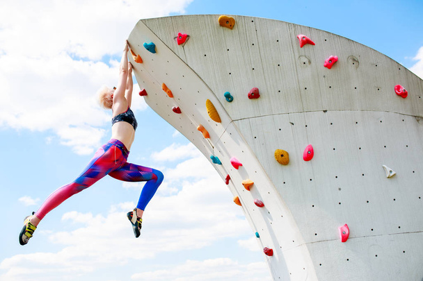 Photo from below of sports woman in leggings hanging on wall for climbing against blue sky - Фото, изображение
