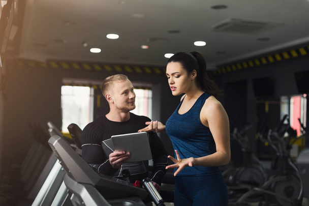 Fitness instructor helps young woman on elliptical trainer. Coach discussing workout plan with girl on treadmill. Cardio workout in gym, healthy lifestyle, copy space - Photo, Image