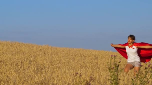 Meisje speelt superheld die dwars over het veld met tarwe onder de blauwe hemel - Video