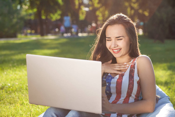 Красива молода жінка, сидячи на бобовій сумці, використовує ноутбук, відпочиваючи на траві в парку на сонці
 - Фото, зображення