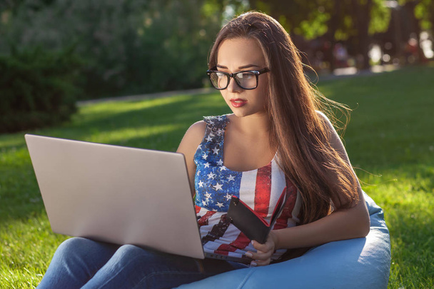 Молодая симпатичная девушка с ноутбуком, сидит на фасолевом пакете в саду или парке, на зеленой траве. Современный образ жизни
 - Фото, изображение