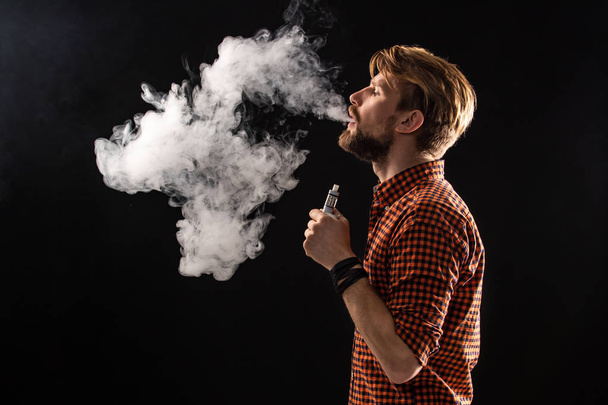 Ένας νεαρός άνδρας με μούσι και ένα κομψό χτένισμα σε ένα πουκάμισο, μια οχιά, ένα δωμάτιο, ένα τσιγάρο, ένα στούντιο, καπνός, το κάπνισμα απόλαυση - Φωτογραφία, εικόνα