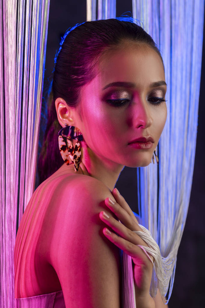 A beautiful glamorous brunette girl with naked shoulders wearing large elegant earrings peeps through a translucent curtain made of white threads. Multicolored artistic stage lighting. Copy space. - Foto, immagini