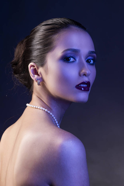 Beautiful brunette girl with naked shoulders and dark red lips make-up, wearing earrings and a pearl necklace, illuminated in blue. Conceptual low key photo. Copy space. Advertising design of jewelry. - Foto, immagini