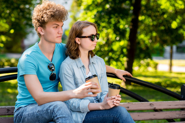 Young beautiful couple of students. Summer in park in nature. Sits on the bench. He holds cups of coffee in his hands. In sunglasses. Enjoys relaxation after school. - 写真・画像