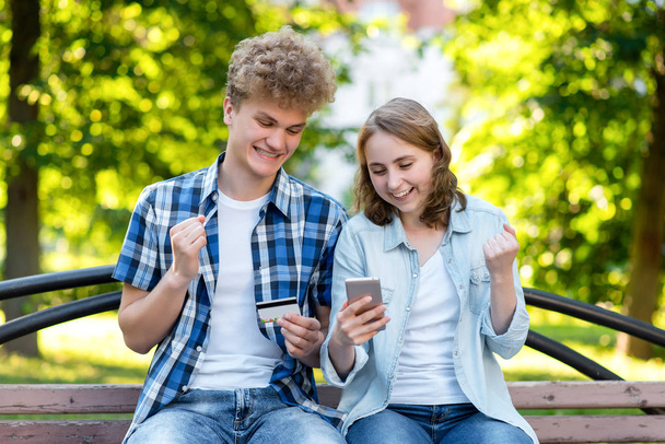 The guy with girl smiles happily. Emotion of winning large sums in the lottery. In his hands holds a smartphone and a credit card. In summer, on a park bench. The concept of success and good luck. - Foto, Bild