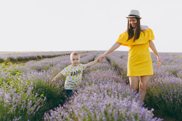 junge Frau in gelbem Kleid zu Fuß auf lila Lavendelblume Wiese Feld Hintergrund, Ruhe, Spaß haben, spielen mit kleinen niedlichen Kind Baby Boy. Mutter, kleiner Sohn. Familientag, Eltern, Kinder - Foto, Bild