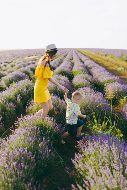 Jonge vrouw in gele jurk lopen op paarse lavendel bloem weide veld achtergrond, rust, plezier, spelen met schattig kind baby jongetje. Moeder, kleine jongen de zoon. Familiedag, ouders, kinderen concept - Foto, afbeelding