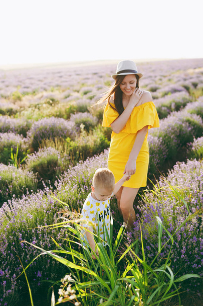 Young woman in yellow dress walk on purple lavender flower meadow field background, rest, have fun, play with little cute child baby boy. Mother, small kid son. Family day, parents, children concept - Fotografie, Obrázek