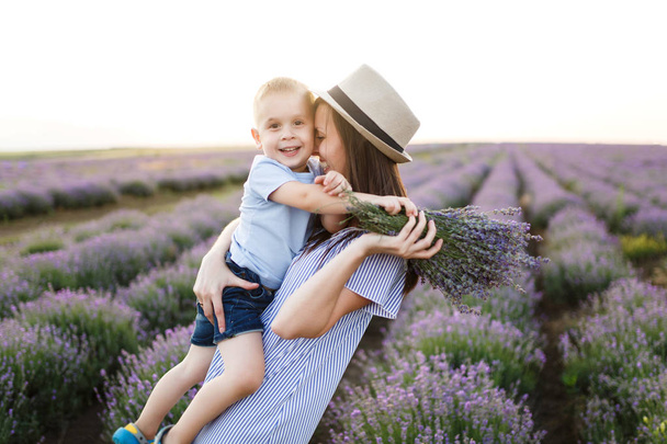 Nuori nainen sininen mekko hattu kävellä violetti laventeli kukka niitty tausta, levätä, pitää hauskaa, leikkiä pieni söpö lapsi poika. Äiti pieni lapsi poika. Perhepäivä, vanhemmat, lasten käsite
 - Valokuva, kuva