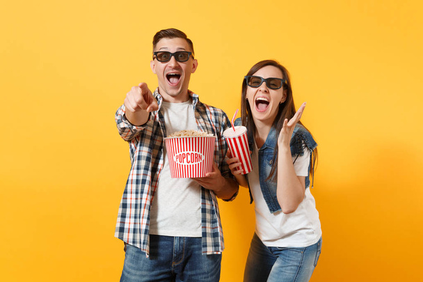 Young laughing couple woman man in 3d glasses watching movie film on date hold bucket of popcorn cup of soda or cola pointing index finger on camera isolated on yellow background. Emotions in cinema - Foto, afbeelding