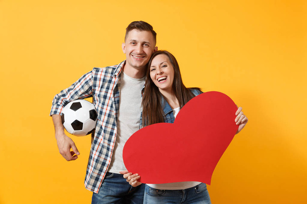 Young happy cheerful couple supporter, woman man, football fans cheer up support team, holding red heart love, soccer ball isolated on yellow background. Sport, family leisure, lifestyle concept - Photo, Image