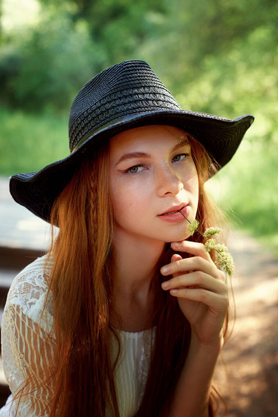 Portrait of a red-haired girl with long hair, in a black wide-brimmed hat.Bites his teeth a blade of grass. Warm sunlight - Foto, immagini