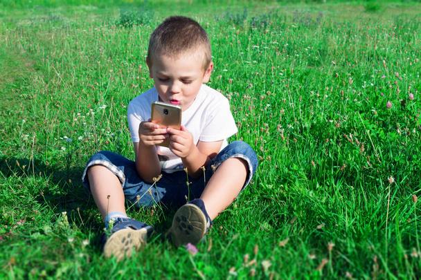 Een jongen is zittend op het gras en het spelen in de mobiele telefoon. - Foto, afbeelding