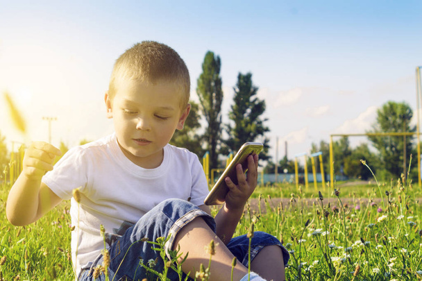 Un bambino è seduto nell'erba sotto il sole con un telefono cellulare in mano
 - Foto, immagini