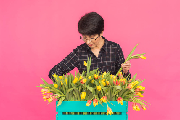 Piękna kobieta w średnim wieku z żółtych tulipanów na różowym tle. Koncepcja florystyki, święta i prezenty - Zdjęcie, obraz