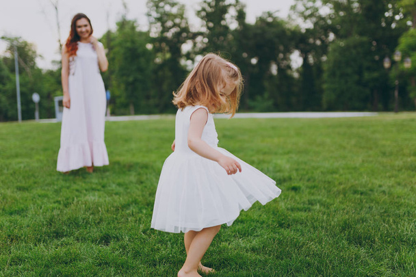 Pretty little cute child baby girl in light dress circling around and have fun on green grass lawn in park. Mother, little kid daughter. Mother's Day, love family, parenthood, childhood concept - 写真・画像