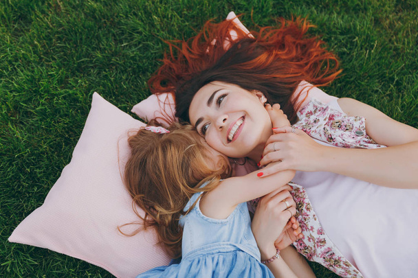 Smiling woman and little cute child baby girl lie on pillows on grass in park hug, embrace and rest, have fun. Mother, little kid daughter. Mother's Day, love family, parenthood, childhood concept - Photo, Image