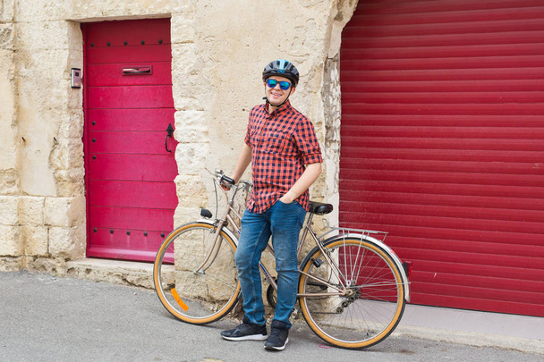 Junger lächelnder Mann mit Helm, rotem Hemd und Sonnenbrille hält Fahrrad im Freien - Foto, Bild