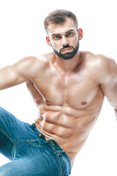 bodybuilder posing. Beautiful sporty guy male power. Fitness muscled in blue jeans. on isolated white background. sits on the floor - Photo, Image