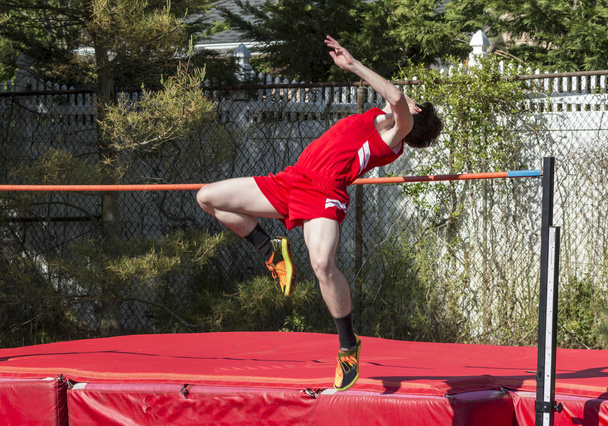 A high school male is over the bar in the high jump with a red uniform on and red matts to land on. - Photo, Image
