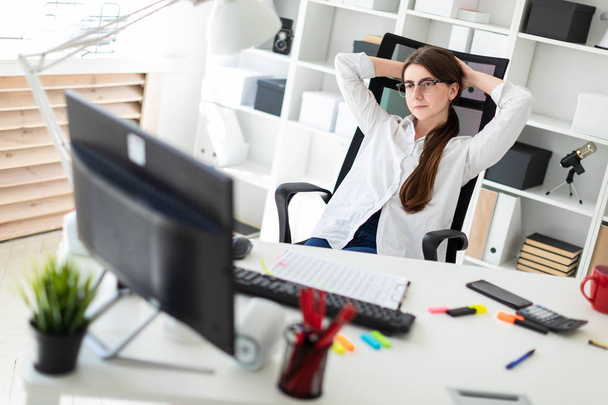 Een jong meisje is zittend aan een tafel in het kantoor, haar handen achter haar hoofd leggen en kijken naar de monitor. - Foto, afbeelding