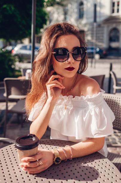 Krásná elegantní mladá žena pití kávy ve venkovní kavárně v létě. Modelka na sobě letní oblečení a doplňky. - Fotografie, Obrázek