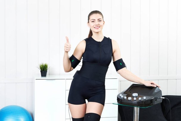 A beautiful young girl stands next to the simulator for practicing electro stimulation of muscles EMS training. The concept of modern fitness using high technologies. - Foto, Bild