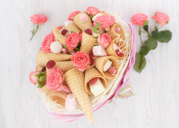 Bouquet consisting of flowers of roses, waffle cones, sweets and marshmallows on a light wooden background - Photo, Image