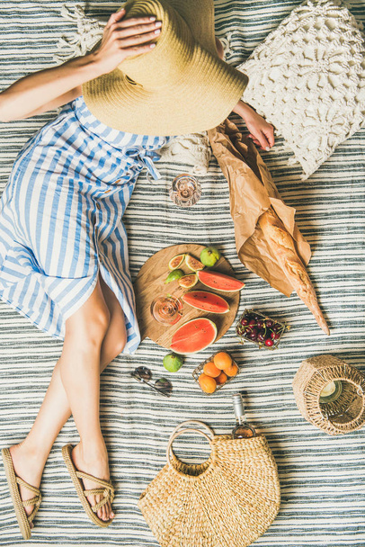 Young woman in striped dress and straw sunhat sitting with rose wine, fresh fruit on board and baguette on blanket - Photo, Image
