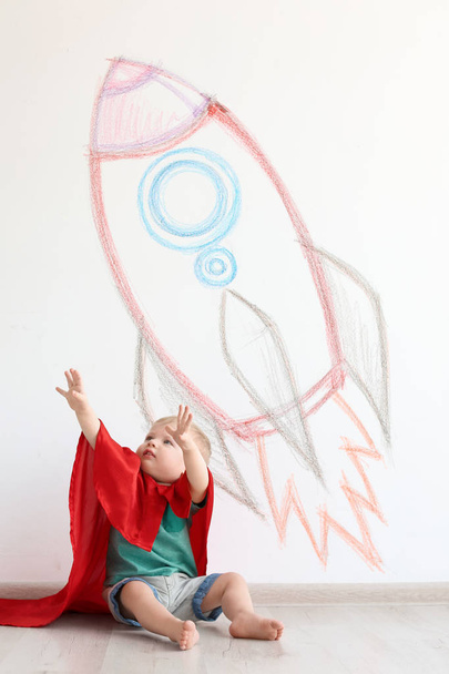 Adorable little child playing astronaut near wall with drawing of spaceship indoors - Фото, изображение