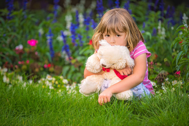 Adorable toddler girl hugging her favourite teddy bear in summer park on beautiful sunny day with yellow flowers in the background - Foto, imagen