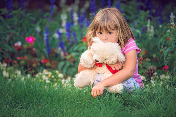 Adorable toddler girl hugging her favourite teddy bear in summer park on beautiful sunny day with yellow flowers in the background - Fotografie, Obrázek