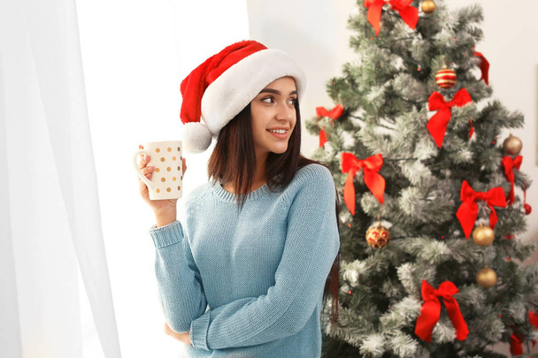 Beautiful young woman in Santa hat with cup of drink near Christmas tree at home - 写真・画像