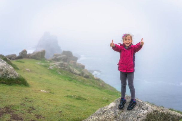 Voittoisa pikkutyttö osoittaa peukalot ylös merkki kiipeämisen jälkeen suuri kallio rannikolla Cornwall, UK
 - Valokuva, kuva