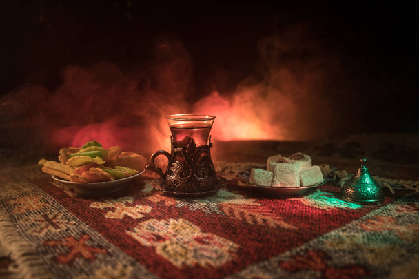 Chá árabe em vidro com lanches orientais em um tapete no fundo escuro com luzes e fumaça. Conceito de chá oriental. Um espaço vazio. Foco seletivo
. - Foto, Imagem