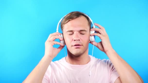 Young man on a colored blue background. portrait. emotions and gestures. 4k, close-up. Slow motion. guy listening to music in white headphones - Кадры, видео