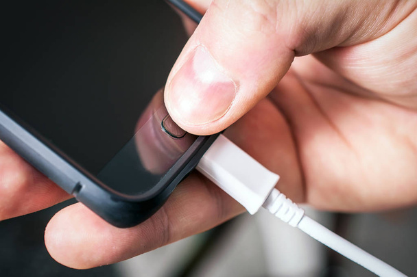 mit einem schwarzen Mobiltelefon, das mit einem weißen USB-Kabel verbunden ist und von einer männlichen Hand gehalten wird - Foto, Bild