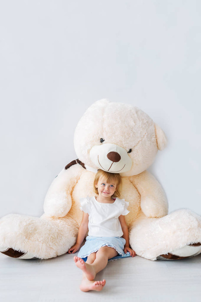 Muotokuva kauniista pikkutytöstä, joka istuu valtavan nallekarhun kanssa lastenhuoneessa. Pystynäkymä
 - Valokuva, kuva