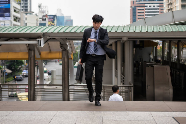 Ανησυχείτε Ασίας επιχειρηματίας τρέχει και να ψάχνει στο ρολόι για να ελέγξετε την ώρα σε βόλτα της πόλης σε ώρα αιχμής. Νεαρός άνδρας αργά για μεταφορά τρένο, εργασία, συνάντηση. Γραφείο ανταγωνισμού ζωή και επιχειρήσεις. - Φωτογραφία, εικόνα