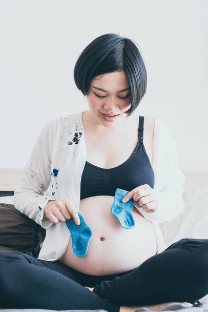 Young beautiful Asian pregnant woman lying in bed and choosing baby socks on bed - selective focus point and vintage  style - Foto, immagini