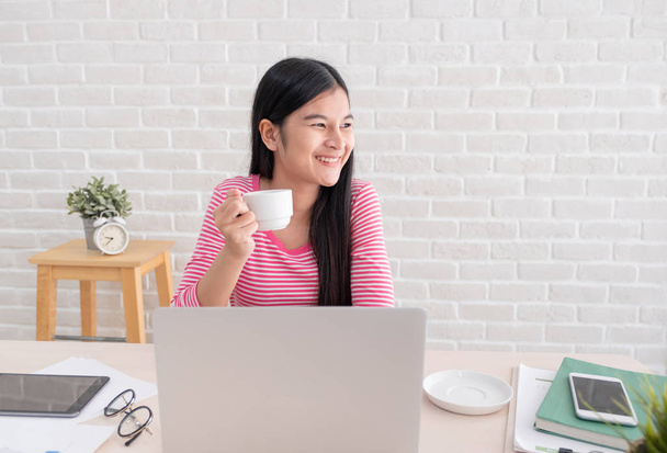 Азиатская женщина фрилансер улыбка и пить кофе с расслаблением эмоций и глядя в окно взять перерыв от ноутбука работы на столе у кирпичной стены в home.working онлайн образ жизни
 - Фото, изображение