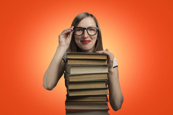 Young girl in a white dress and glasses put her hands on a stack of books on an orange background. Concept of a young student, student and education. - Photo, Image