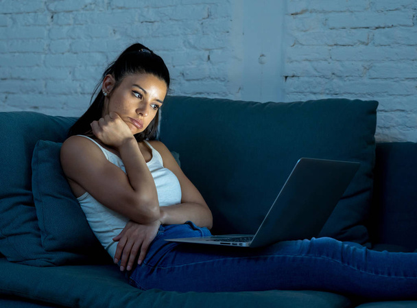 young beautiful hispanic internet addict woman in casual clothes sitting on the couch working bored and tired with laptop computer late at night in dark room light exhausted from work. - Zdjęcie, obraz