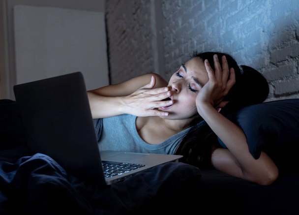 young beautiful hispanic internet addict woman in pajamas on bed at home bedroom working bored and tired with laptop computer late at night in dark room light yawning exhausted falling sleep. - Foto, immagini