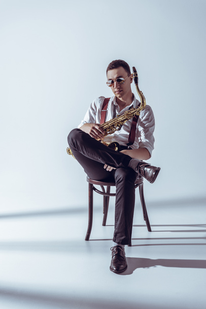 Stilvoller junger Performer mit Sonnenbrille sitzt auf Stuhl und hält Saxophon auf grau - Foto, Bild